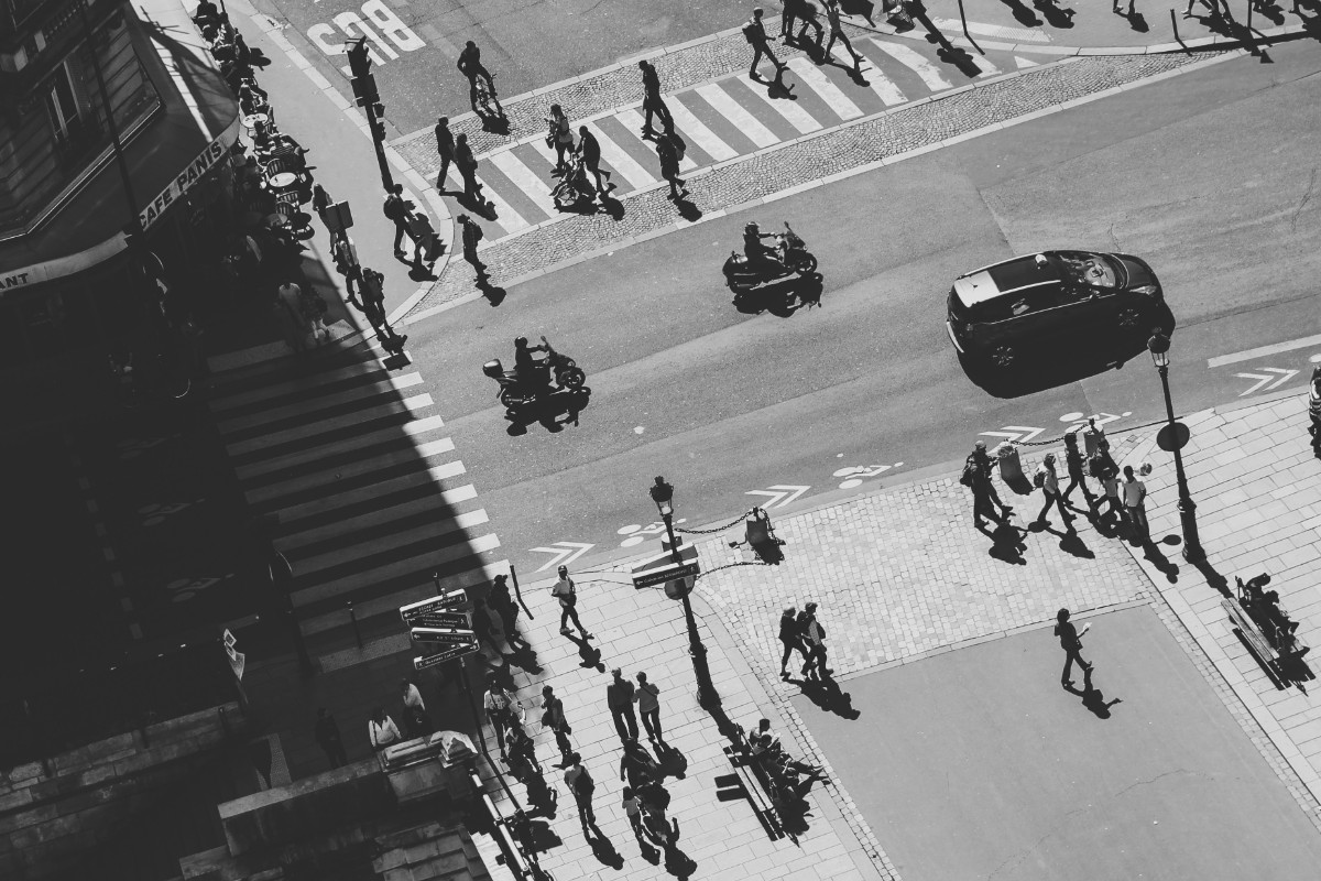 Aerial view of a busy city intersection with numerous people crossing the street and a few vehicles passing by. Shadows are visible in the black and white image.