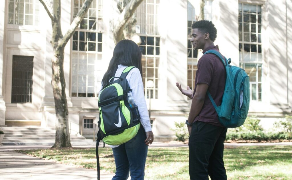 Two students with backpacks stand and talk outside a building with large windows.