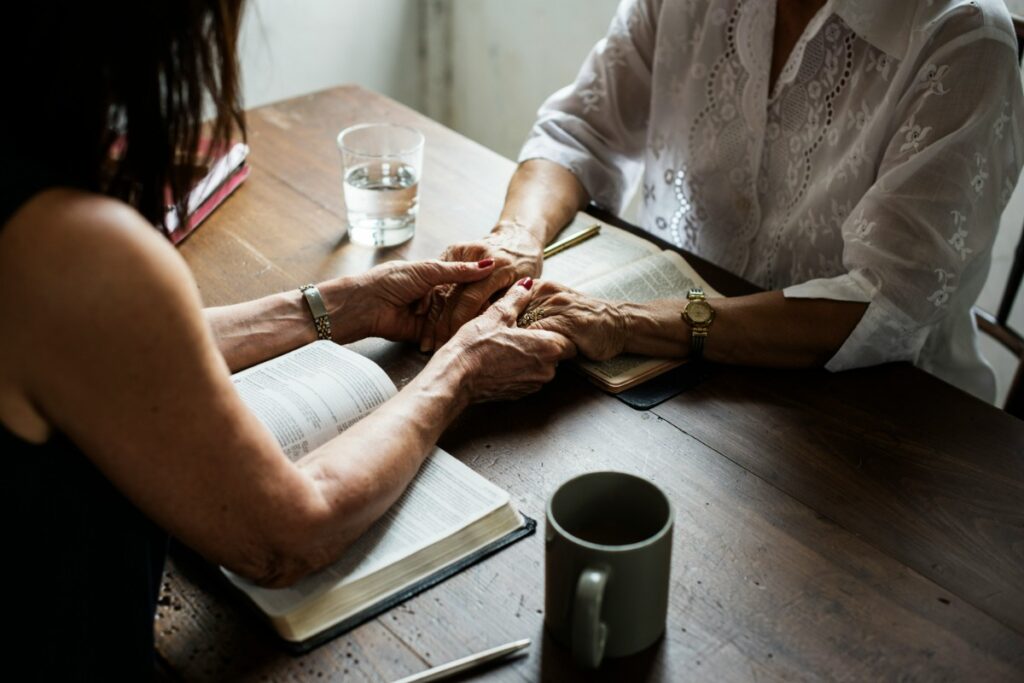 Two people sit at a table holding hands over open books. A notebook, pen, glass of water, and mug are also on the table.