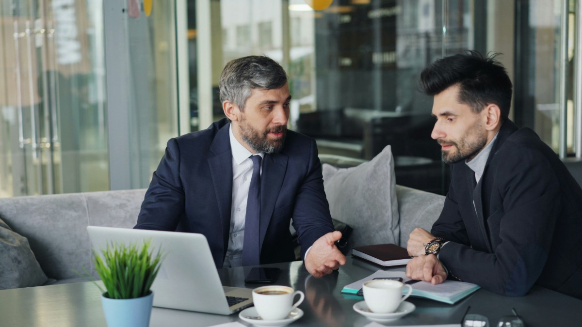 Two men in suits sit at a table with a laptop, coffee cups, and notebooks. They are engaged in conversation in a modern, light-filled office setting.