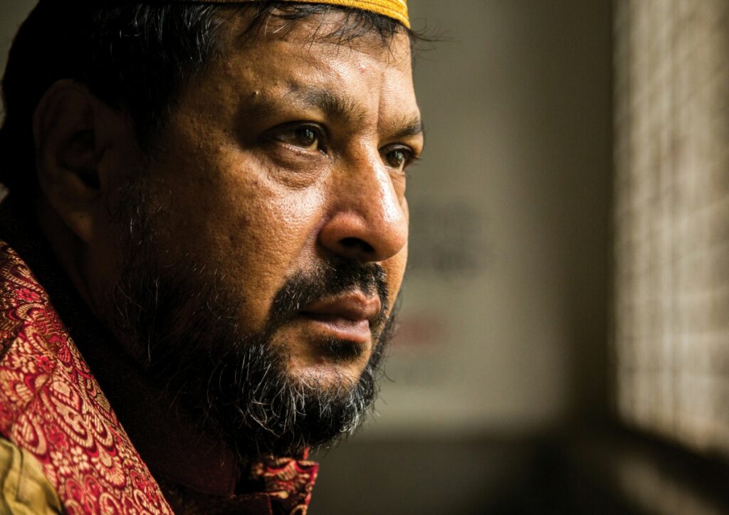 A man with a beard wearing a yellow cap and red patterned garment looks thoughtfully out of a window.
