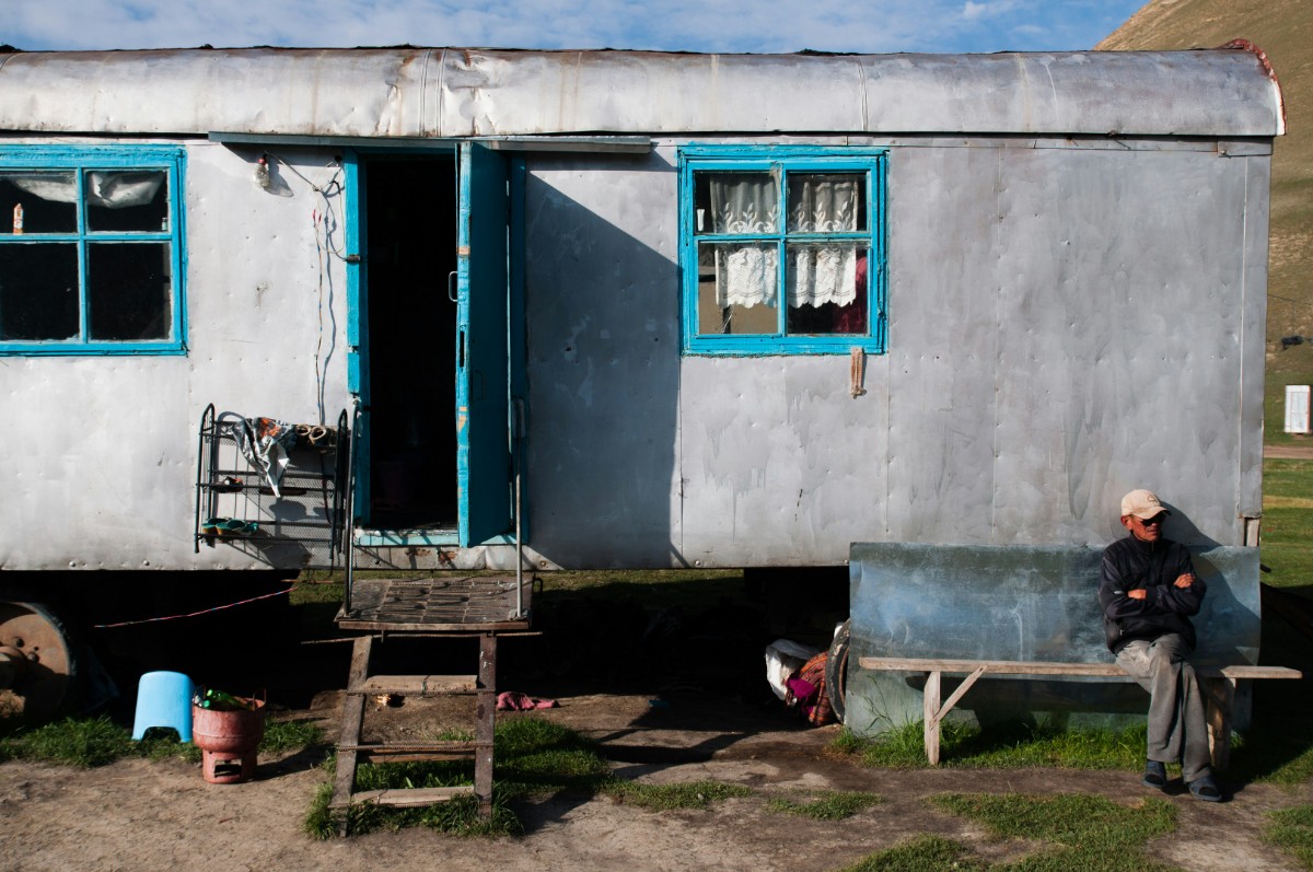 Old trailer with blue-framed windows, open door, and steps in front. A person sits on a bench to the right, under a bright sky.
