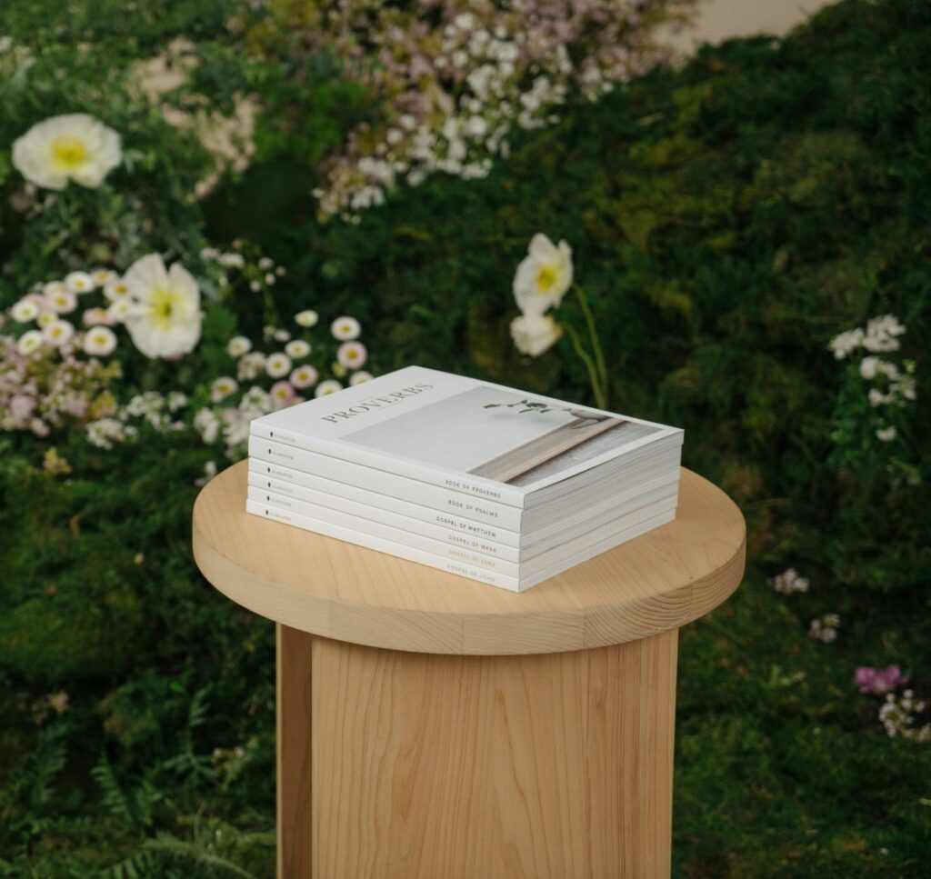 A stack of white books on a wooden pedestal, set against a background of green foliage and white flowers.