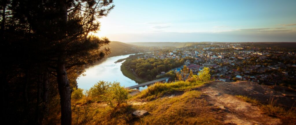 Aerial view of a river winding through a lush landscape with a town on the right. A tree is in the foreground, and the sky is clear with a warm sunset glow.