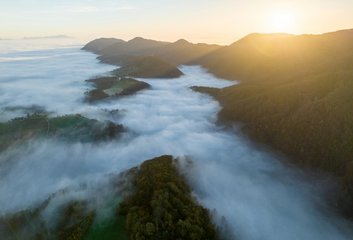 Aerial view of sunlit mountains partially covered by a sea of clouds, with sunlight breaking through.