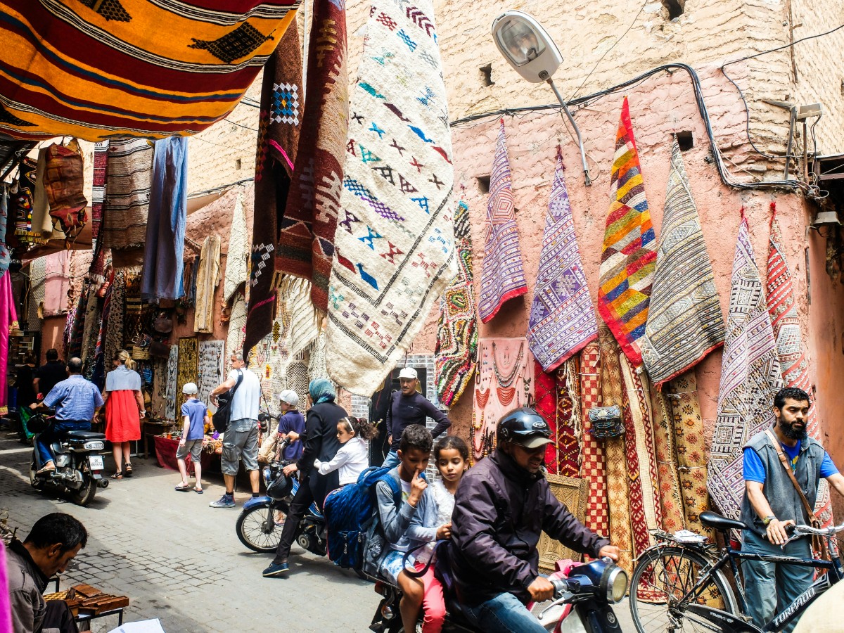 People walking and biking in a narrow, busy street lined with colorful rugs hanging on walls.