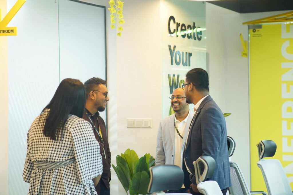 Four people standing and conversing in an office conference room with yellow and white decor.
