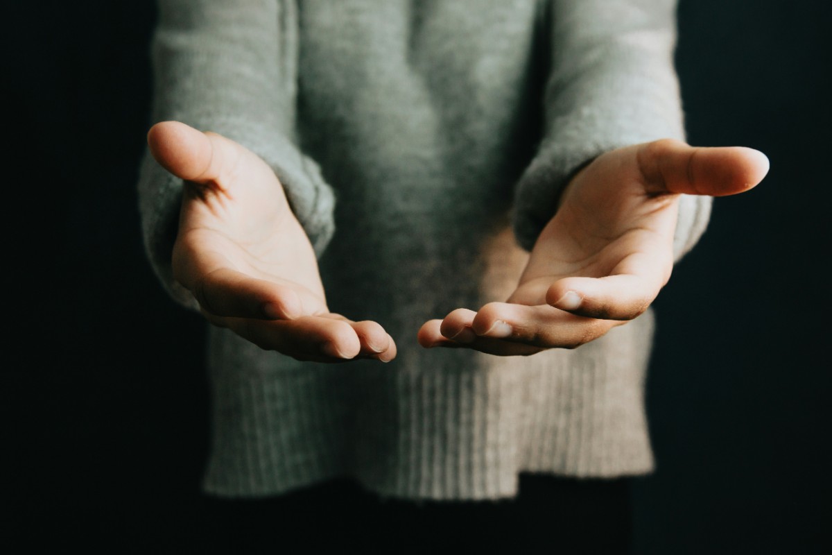 A person wearing a gray sweater holds out both hands with palms facing up against a dark background.