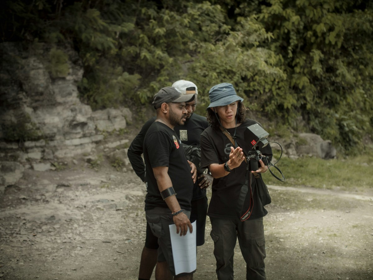 Three individuals stand outdoors reviewing footage on a camera. They are surrounded by trees and rocky terrain.