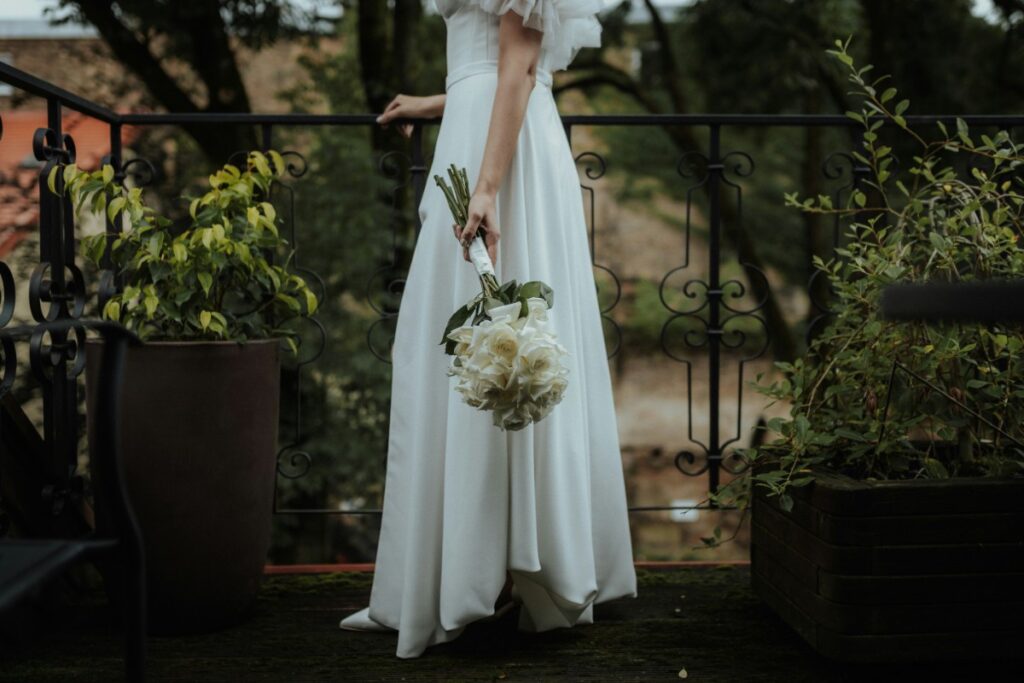 Bride in a white dress holding a bouquet of white flowers, standing on a balcony surrounded by plants.