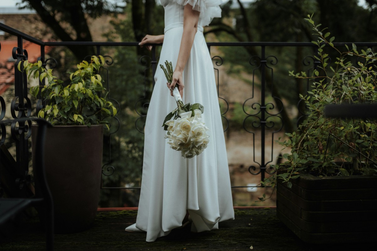 Bride in a white dress holding a bouquet of white flowers, standing on a balcony surrounded by plants.