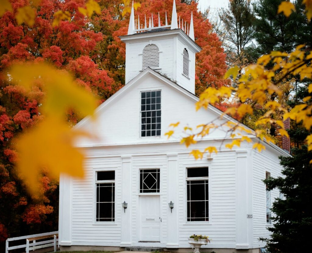 A small white church with a steeple stands amidst vibrant autumn trees with red and yellow leaves in the foreground.