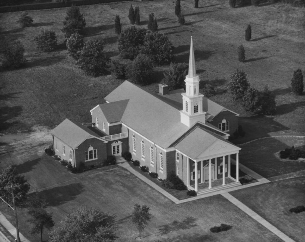 Aerial view of a church with a tall steeple and columns, surrounded by grass and trees, on a clear day.