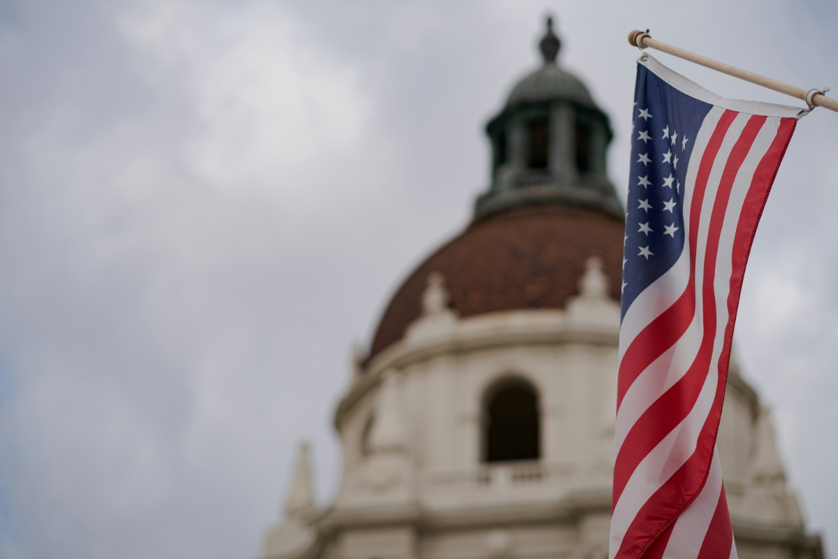 An American flag waves with a blurred dome of a classical building in the background under a cloudy sky.