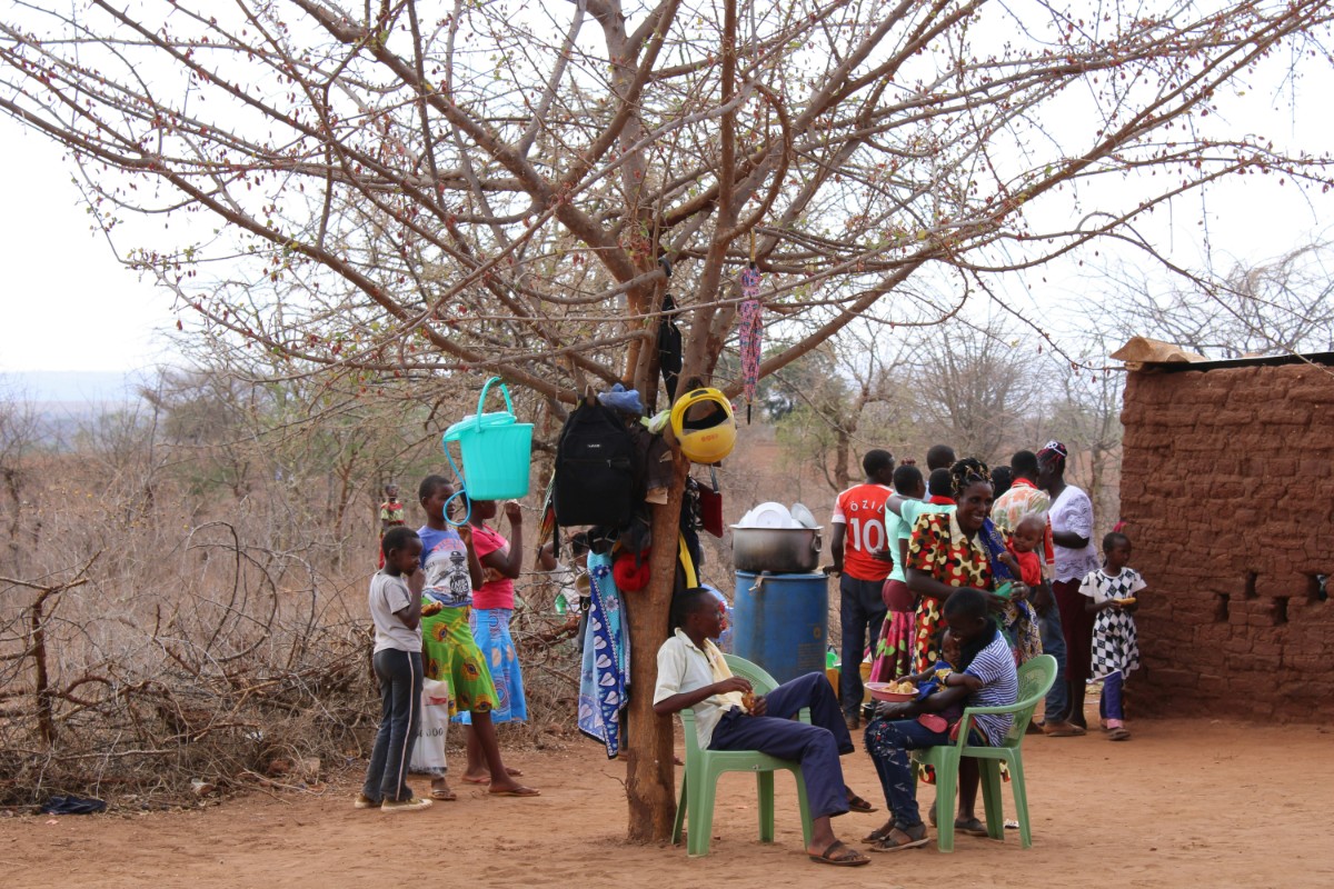 Group of children and adults gather near a leafless tree and a mud-brick building in a dry area. Some are seated, while others stand or tend to household items under the tree.