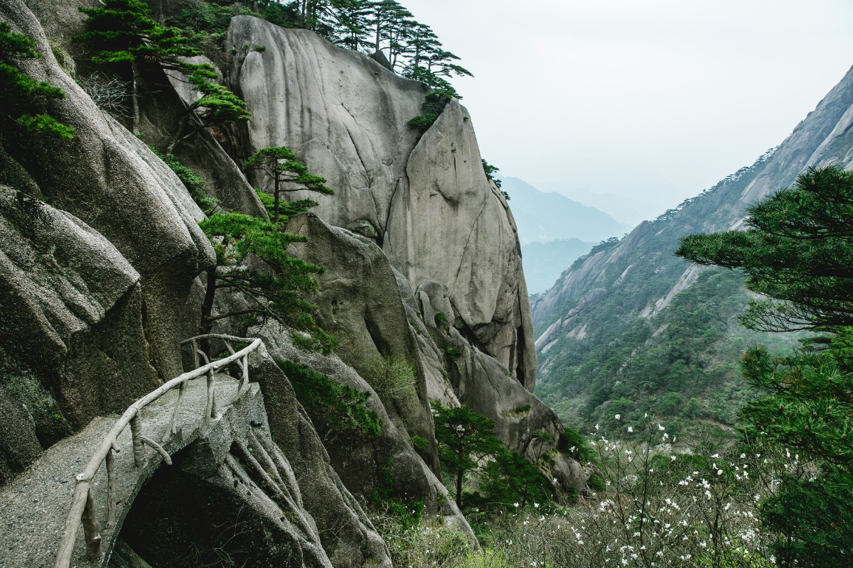 Stone pathway alongside steep rocky cliffs with scattered greenery and misty mountains in the background.