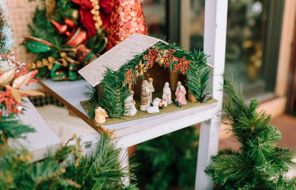 A small Nativity scene with figurines is displayed on a shelf, surrounded by green foliage and red decorations.