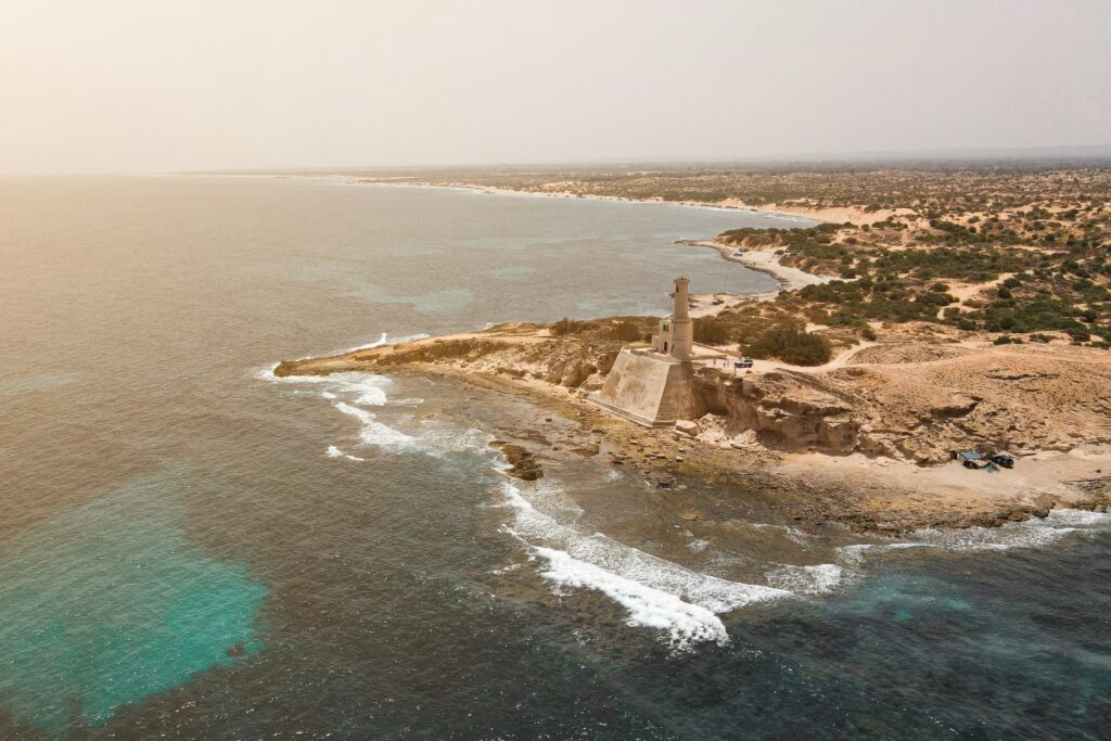 Aerial view of a coastal landscape with a lighthouse on a rocky point. The sea is calm, with waves gently hitting the shore, and the land is dotted with sparse vegetation.
