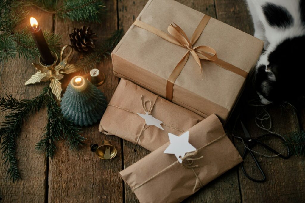 Brown paper-wrapped gifts with star tags and a bow on a wooden surface, surrounded by a candle, pine branches, and a glimpse of a cat.