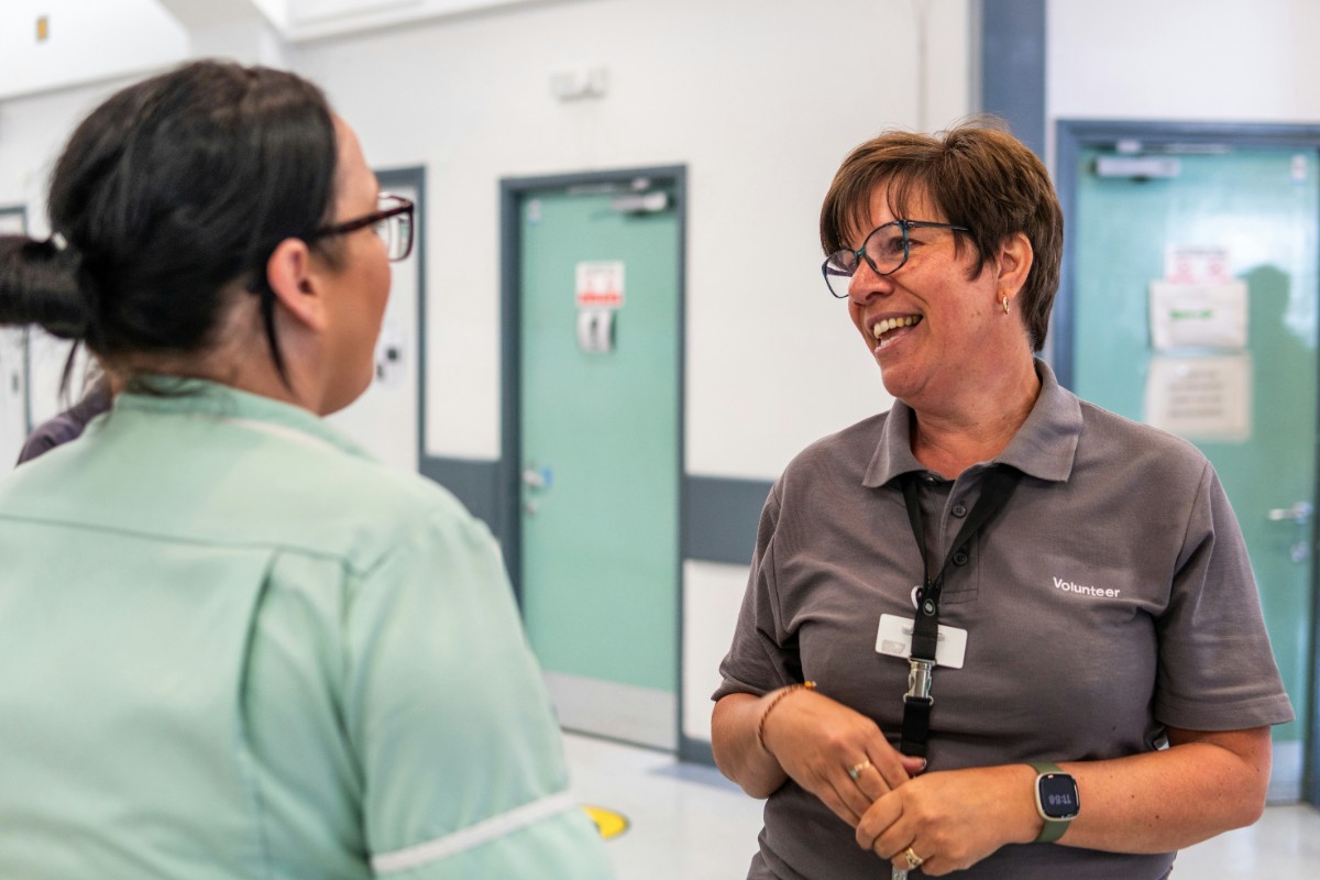 Two women conversing in a hallway. One wears a lanyard and ID badge, smiling, while the other is seen from behind with glasses and a light green uniform.