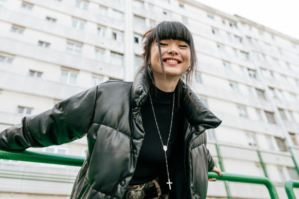 Person smiling in front of a tall building, wearing a black puffer jacket and a necklace with a cross.