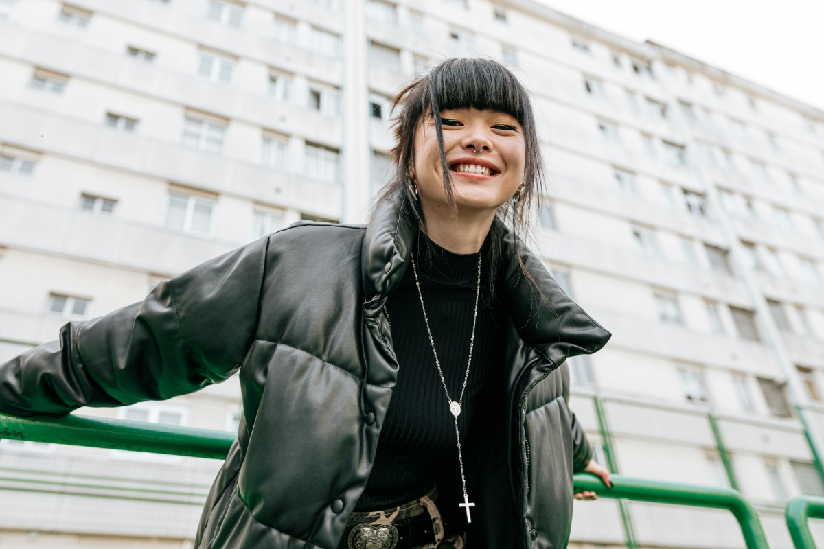 Person smiling in front of a tall building, wearing a black puffer jacket and a necklace with a cross.