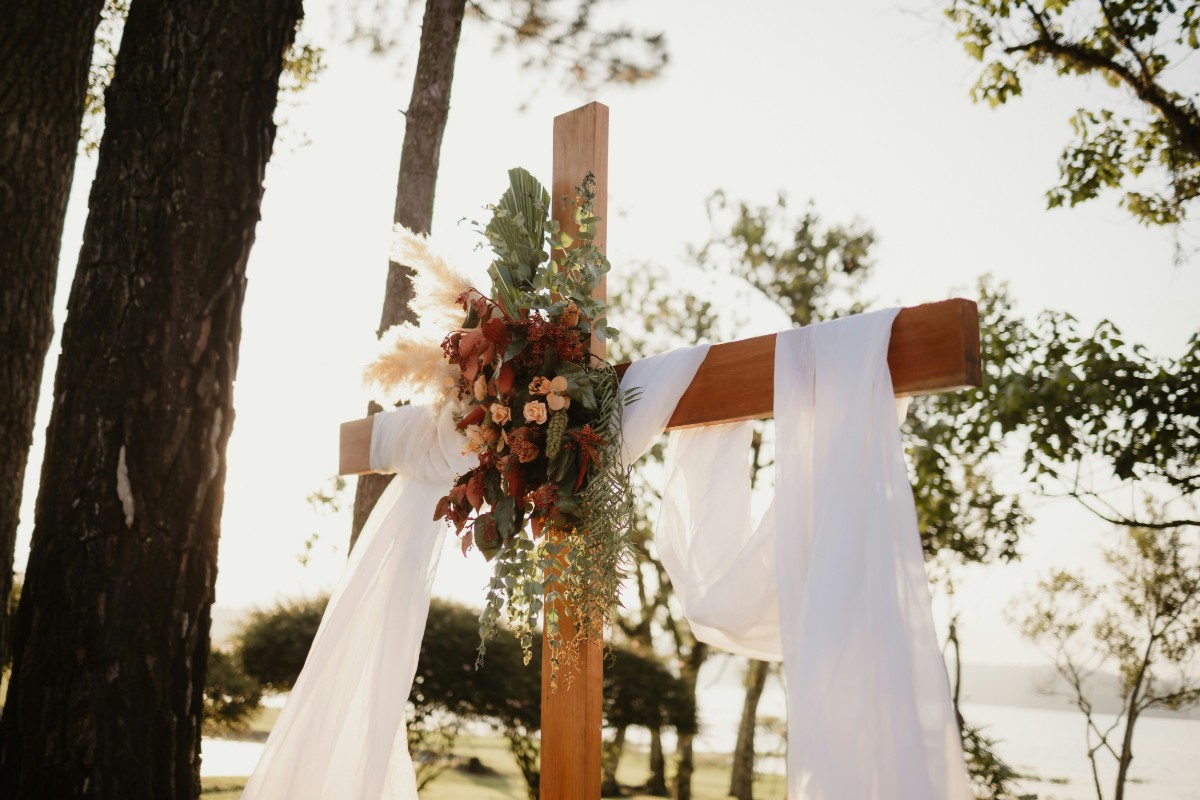 A wooden cross decorated with white fabric and flowers stands outdoors surrounded by trees.