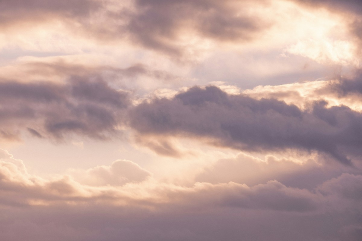 Cloudy sky with soft, muted colors during sunset or sunrise, featuring layers of dark and light clouds partially obscuring the light.