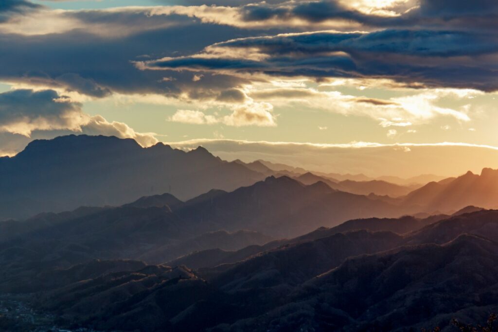 Mountain range at sunset with layers of peaks and valleys, under a partly cloudy sky.