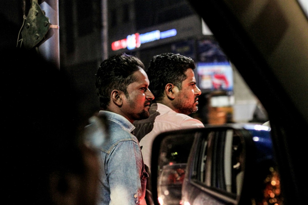 Two people standing beside a vehicle at night, visible in the side mirror, with a blurred city background.