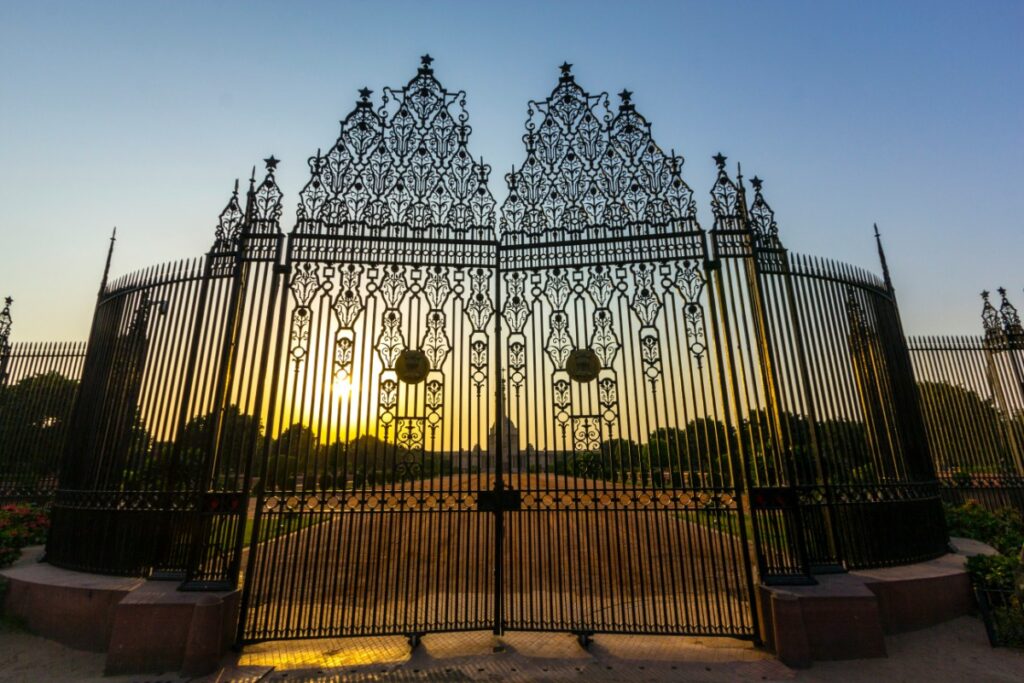 Ornate metal gates with intricate designs stand open, framing a pathway. The sun sets in the background, casting a warm glow over the scene.