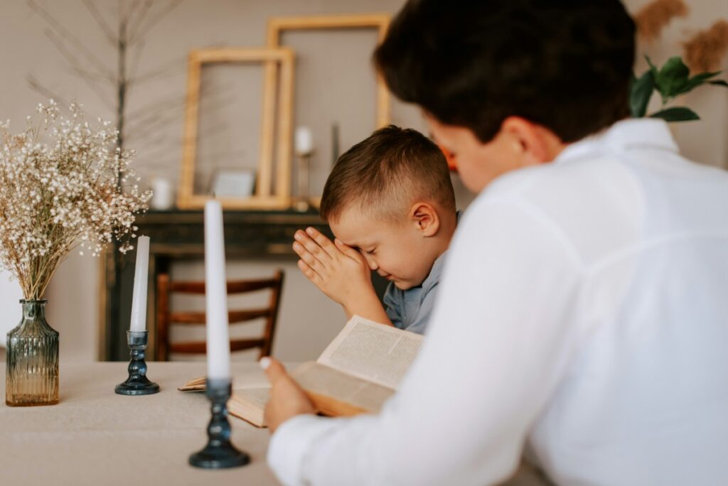 A child and an adult sit at a table with a book. The child has hands clasped in front of their face, and there are two lit candles and a vase with flowers on the table.