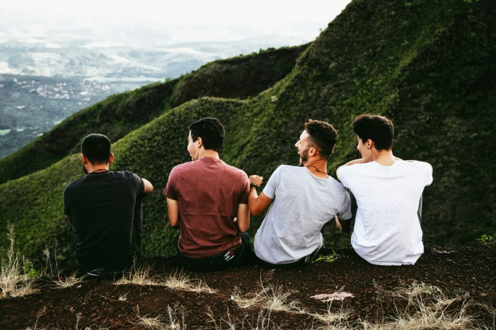 Four people sitting on a grassy hill, facing away, overlooking a scenic landscape with green hills and a distant view.