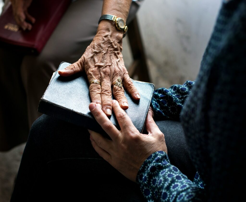Two hands gently touching a closed book. One hand is elderly and adorned with rings and a wristwatch, while the other is younger, partially obscured by a patterned sleeve.