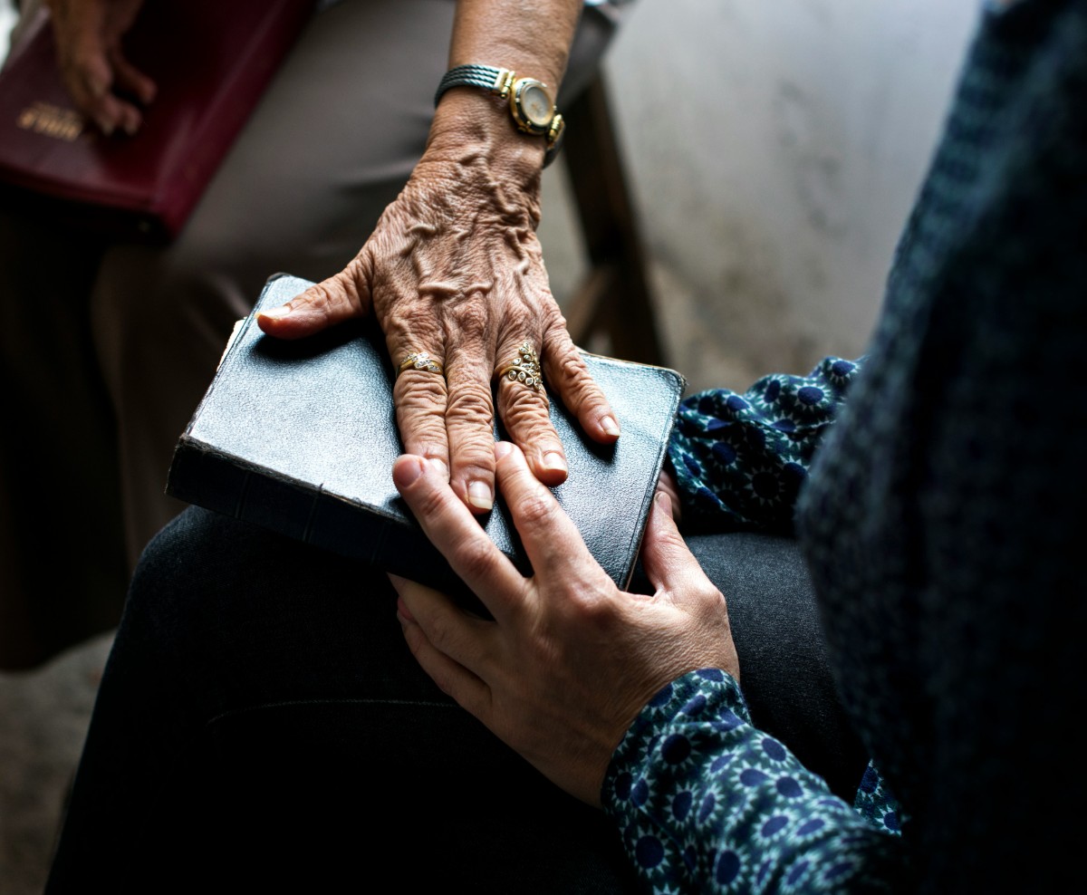 Two hands gently touching a closed book. One hand is elderly and adorned with rings and a wristwatch, while the other is younger, partially obscured by a patterned sleeve.