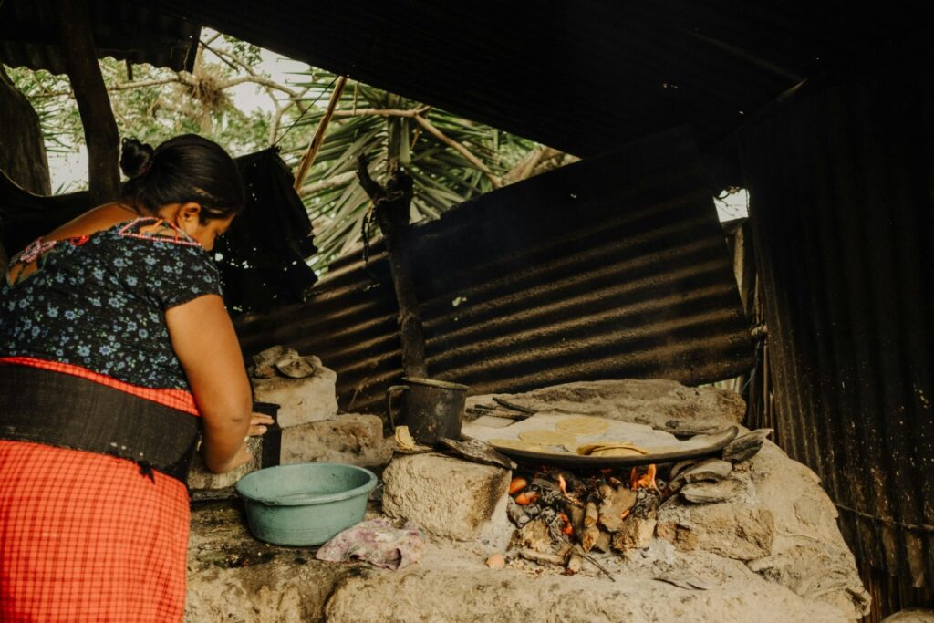 A woman in a patterned top and red skirt cooks on an outdoor stone stove with a pot and a flatbread. A blue bowl is on the side. Trees and corrugated metal walls are in the background.