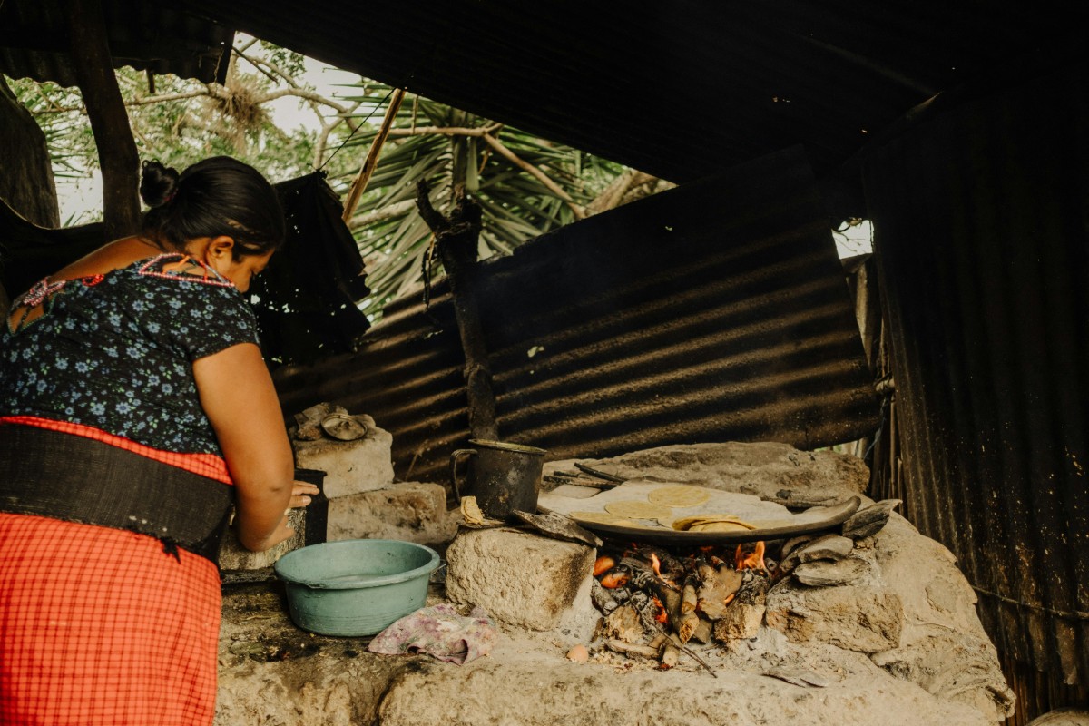 A woman in a patterned top and red skirt cooks on an outdoor stone stove with a pot and a flatbread. A blue bowl is on the side. Trees and corrugated metal walls are in the background.