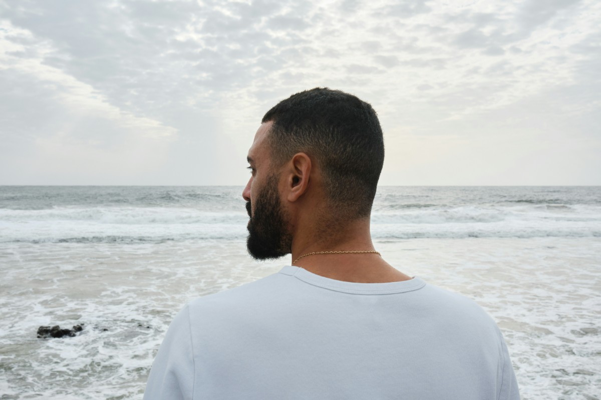 Man in a white shirt standing on the beach, looking at the ocean under a cloudy sky.