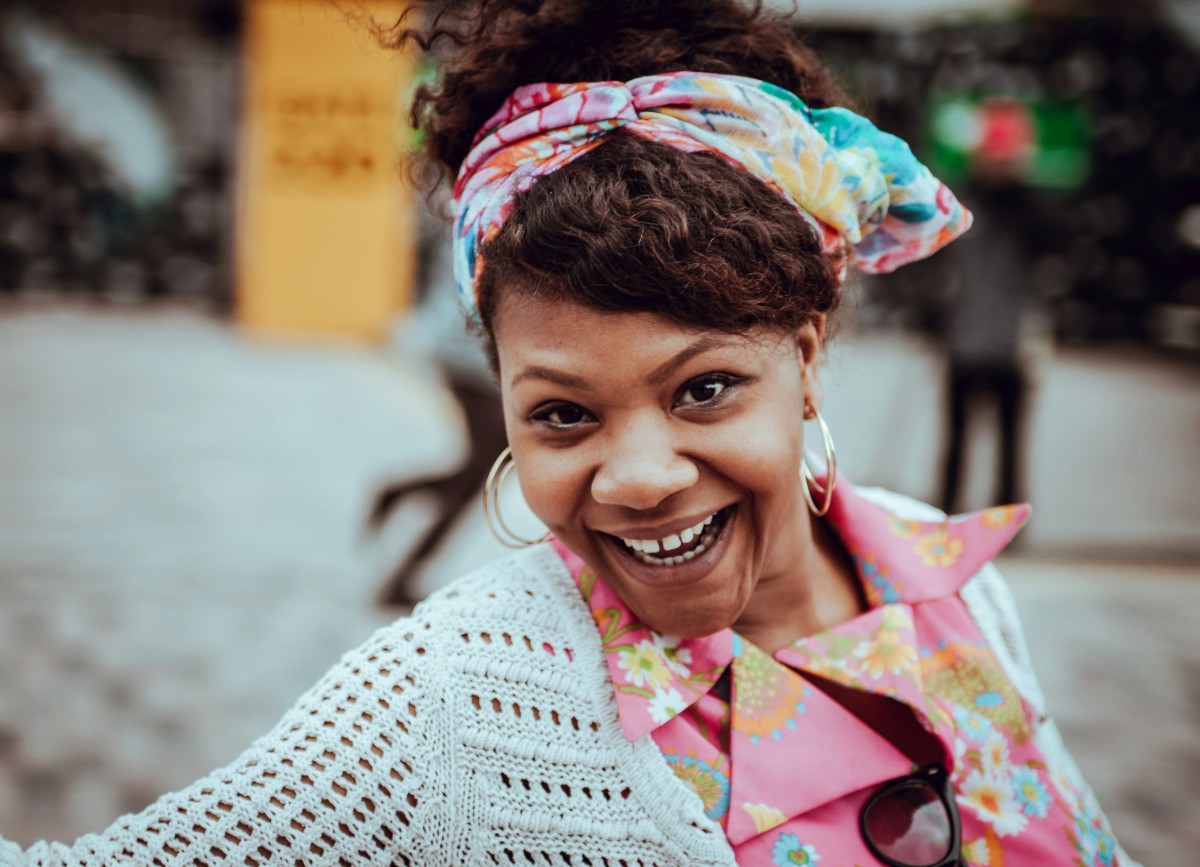 A smiling woman wearing a colorful headscarf and pink shirt poses outdoors.