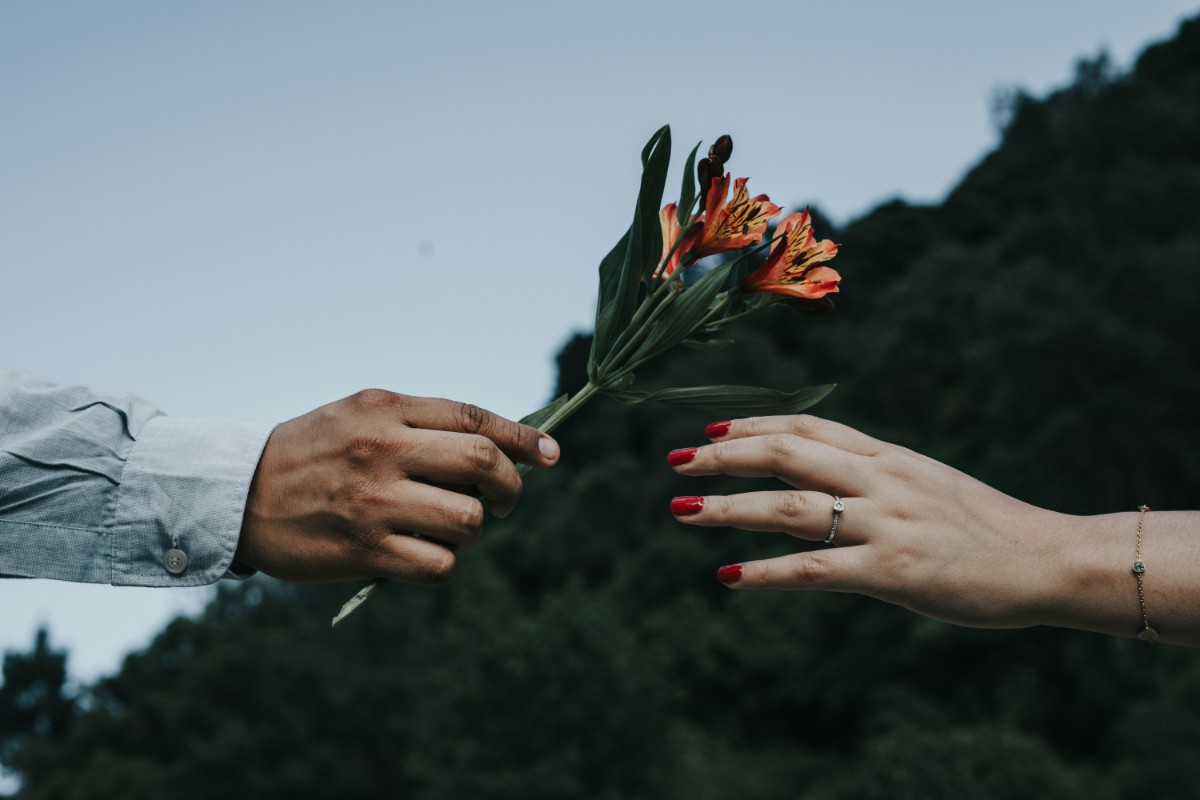 One hand offers a small bouquet of orange flowers to another hand with red-painted nails against a blurred natural background.