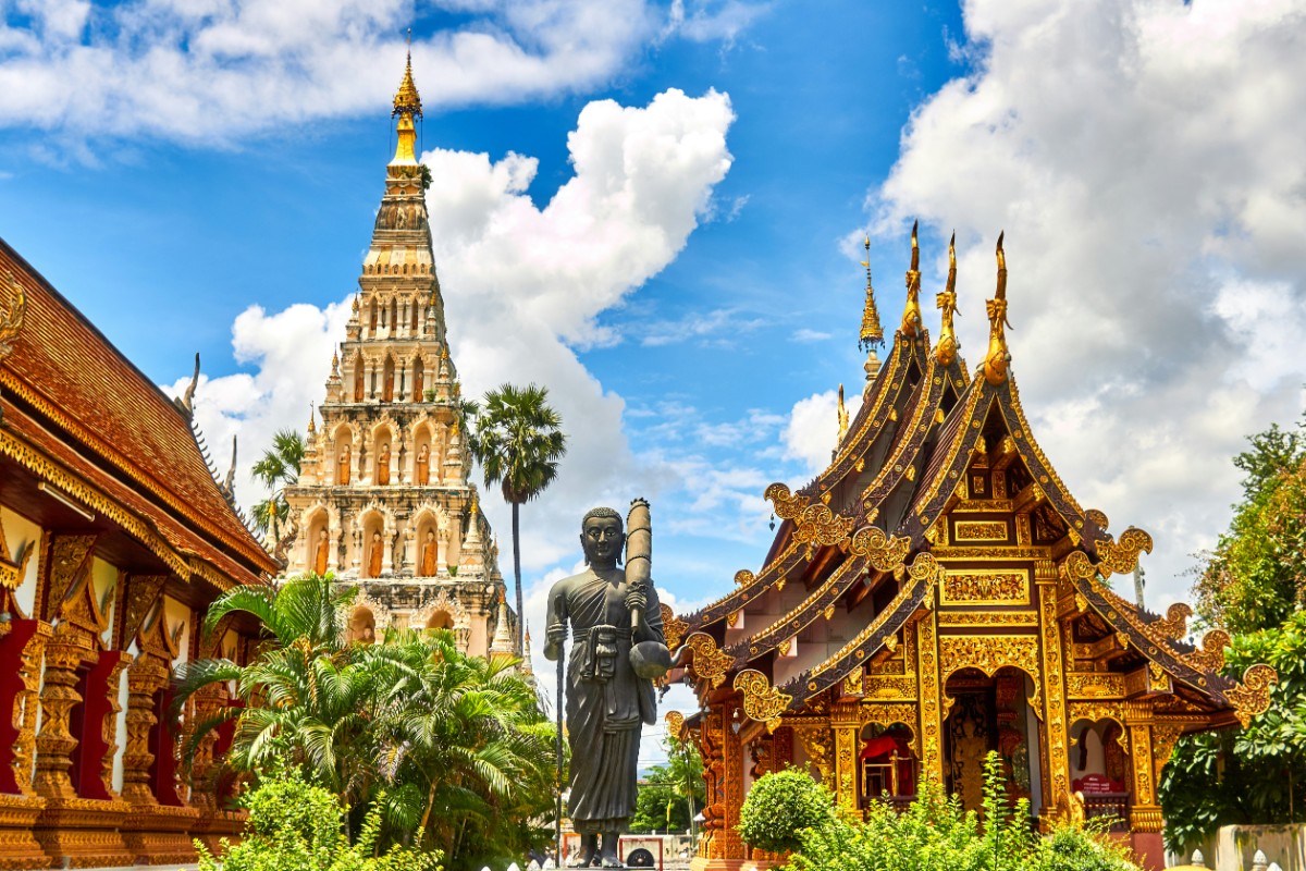 A temple complex featuring a tall pagoda, a statue, and an ornate building with gold details under a blue sky with clouds.