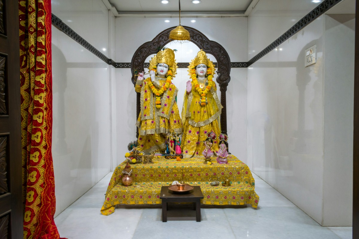 Statues of deities adorned in yellow garments are displayed on a decorated platform, surrounded by small offerings, inside a white-walled room with a decorative arch.