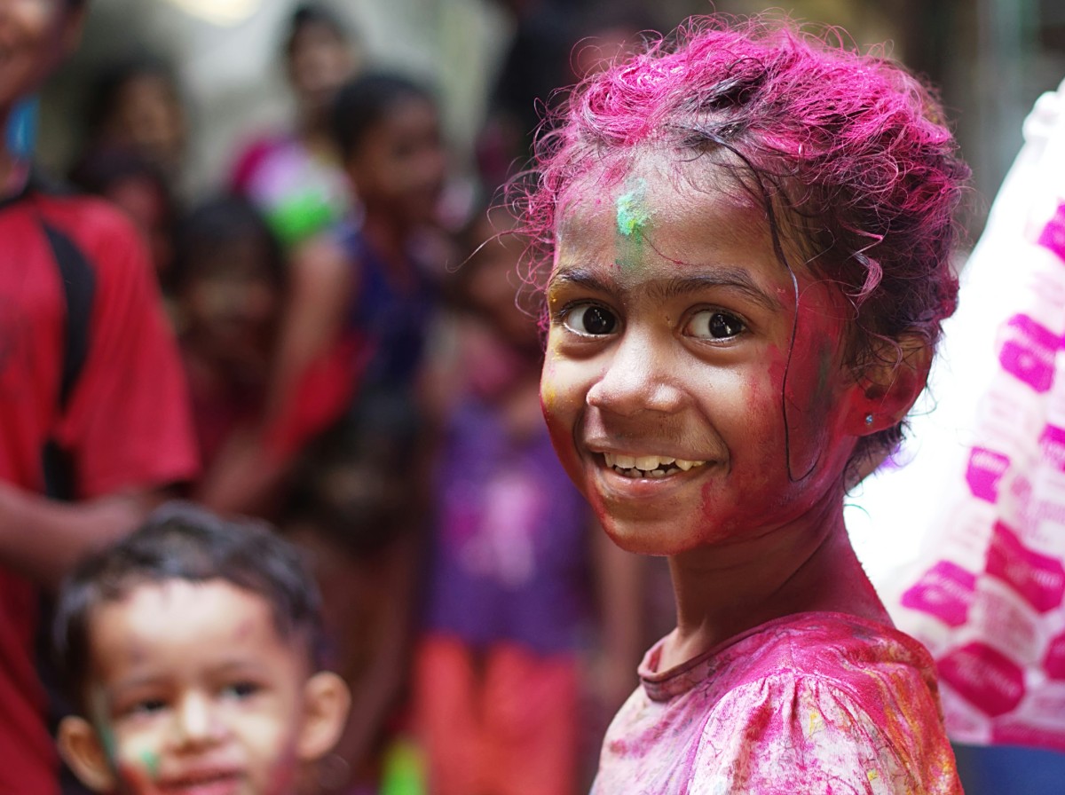 A child smiles, covered in colorful powders, with people in the blurred background, celebrating a festival.
