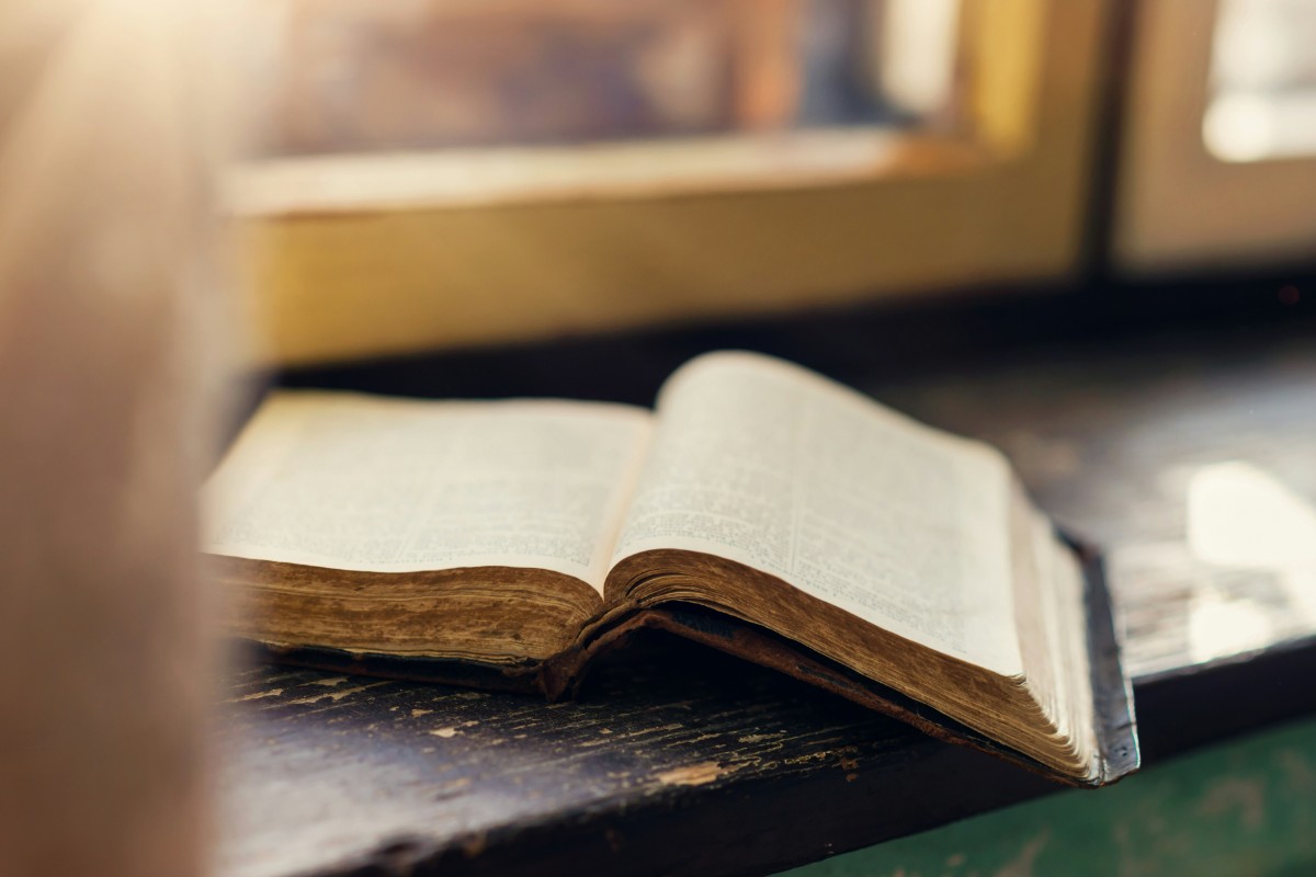 An open book with yellowed pages lies on a wooden windowsill, illuminated by soft natural light.