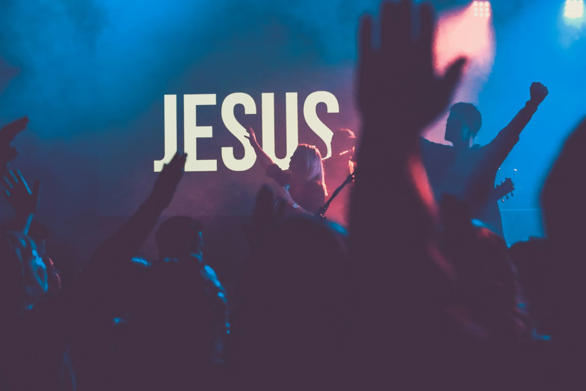 A crowd raises hands in front of a stage with JESUS displayed on a screen, bathed in purple and blue lighting.