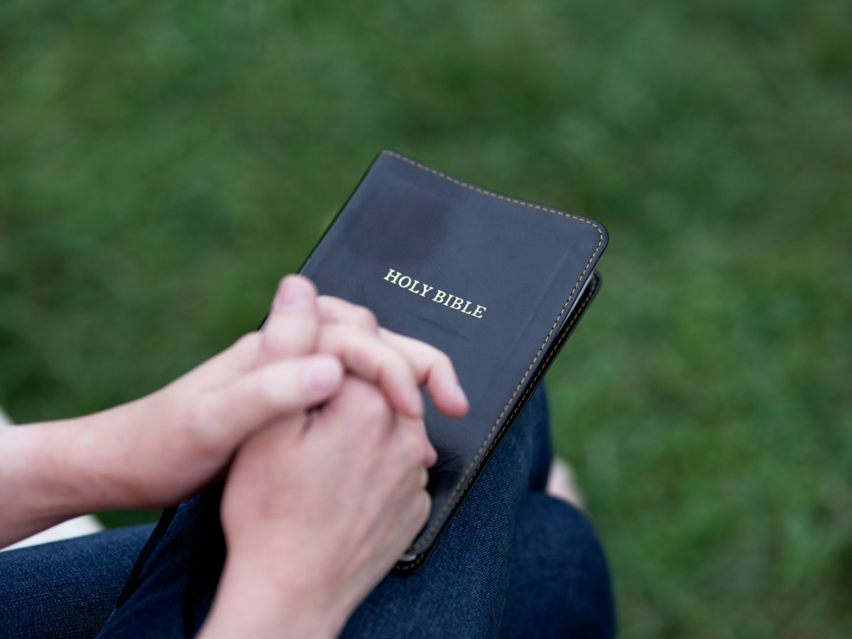 Hands clasped in prayer holding a closed Holy Bible, resting on someones lap with a green grass background.