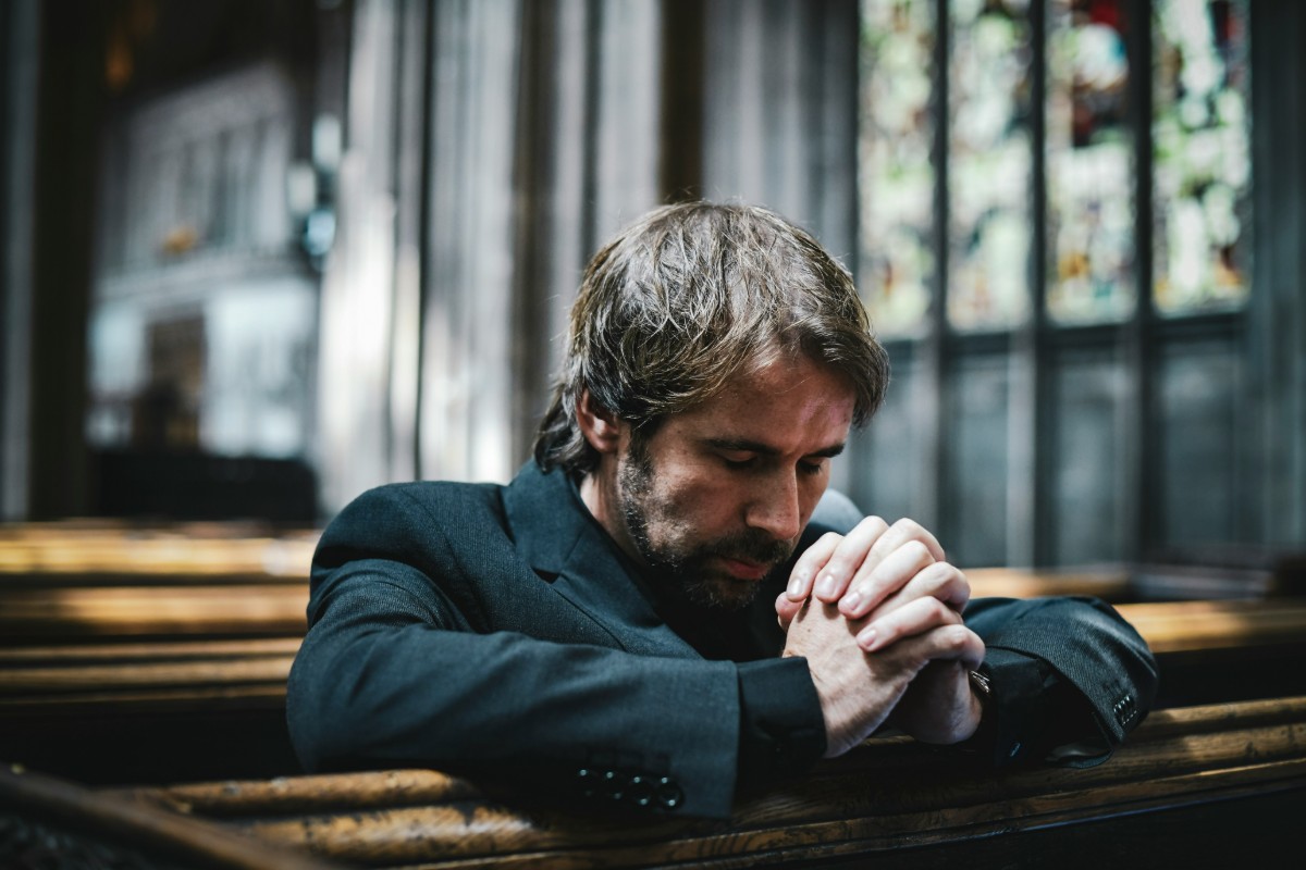 A man with closed eyes and clasped hands sits in a church pew, appearing contemplative or in prayer.