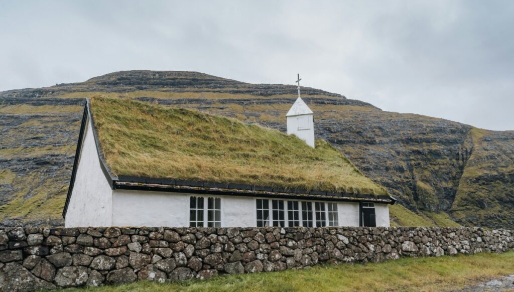 A small white church with a grass-covered roof and a steeple, set against a backdrop of grassy hills and cloudy sky. Stone wall in front.