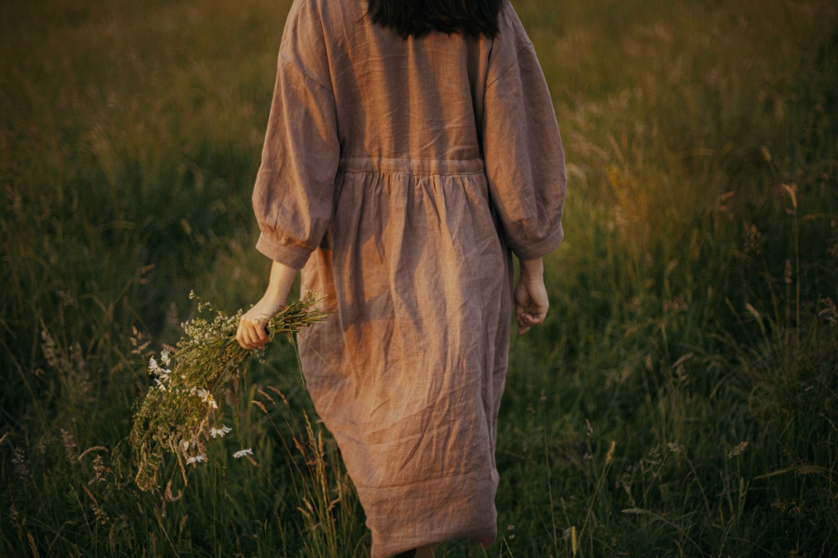 Person in a brown dress walking through a grassy field, holding small white flowers in one hand.