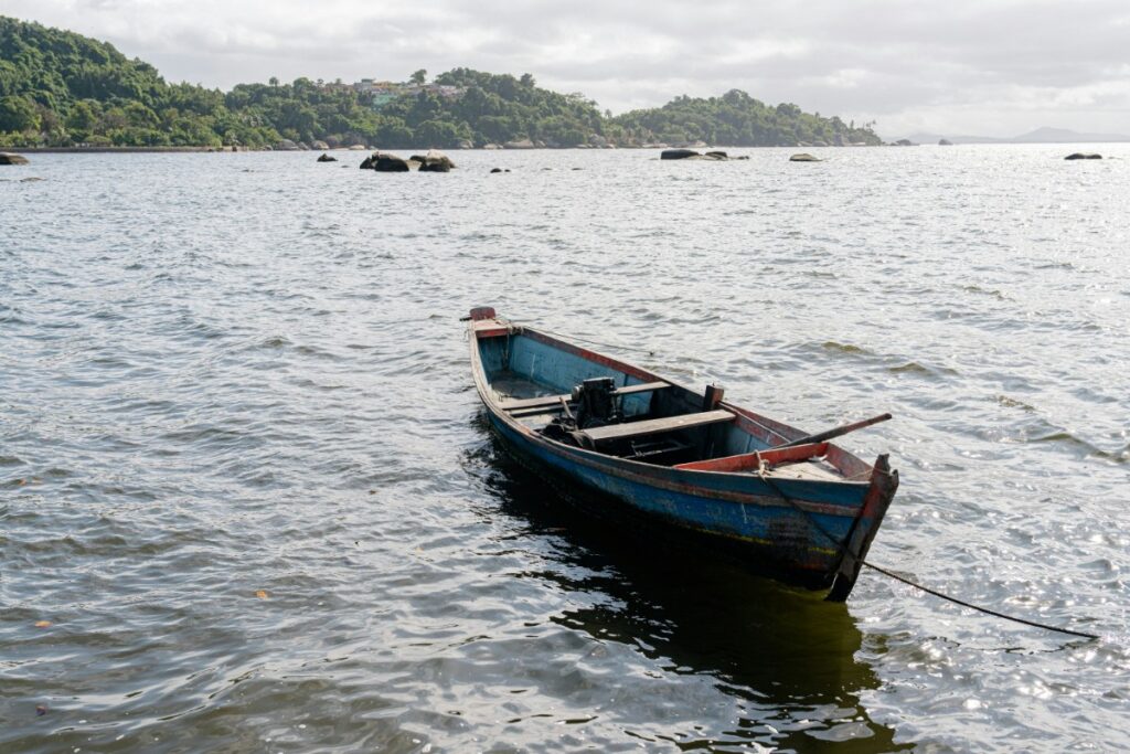 A small, empty wooden boat floats on calm water near a forested shoreline under a cloudy sky.